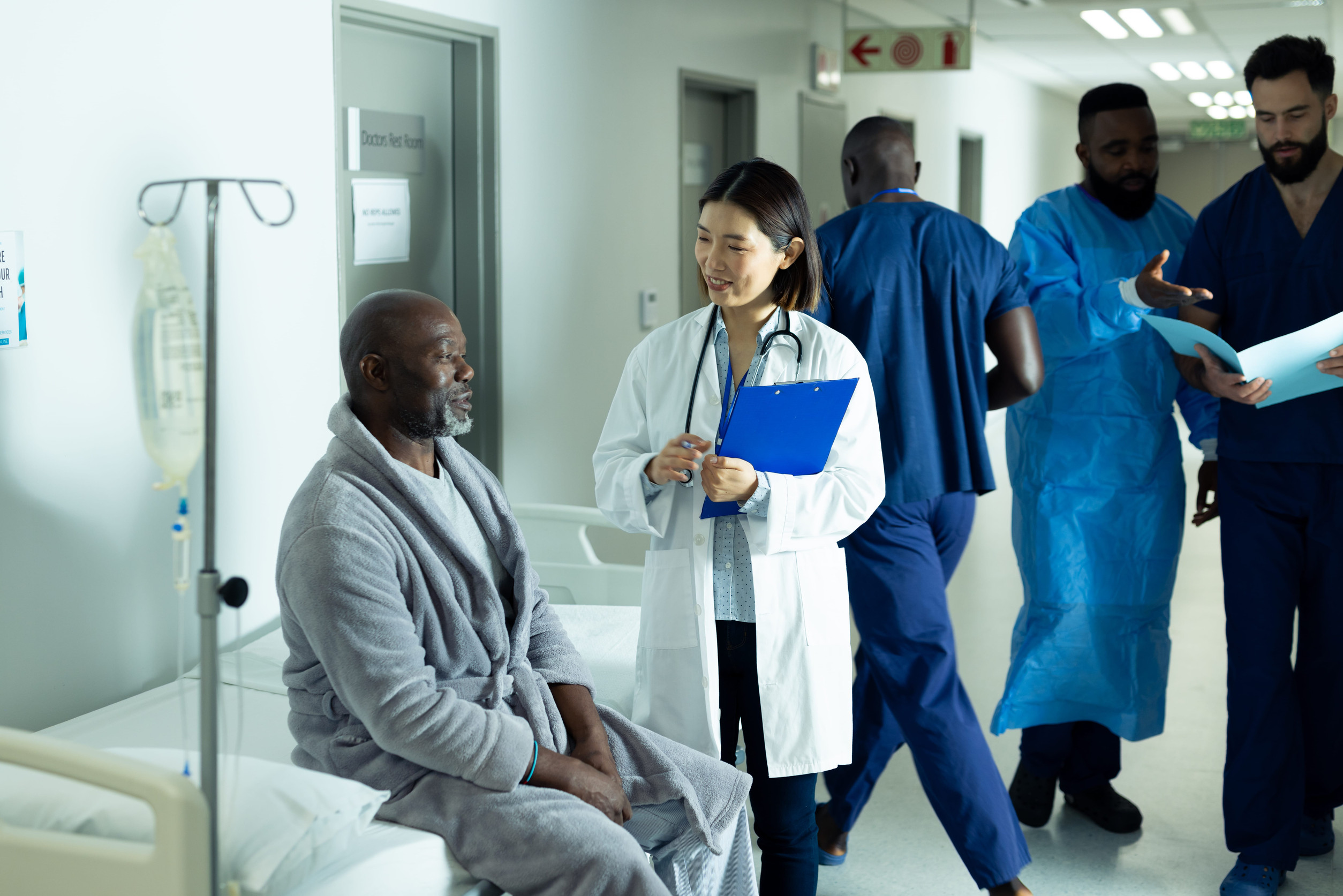 Doctor discussing care with a patient seated beside a hospital bed while medical staff work in the background, representing coordinated healthcare, patient safety, and clinical teamwork.